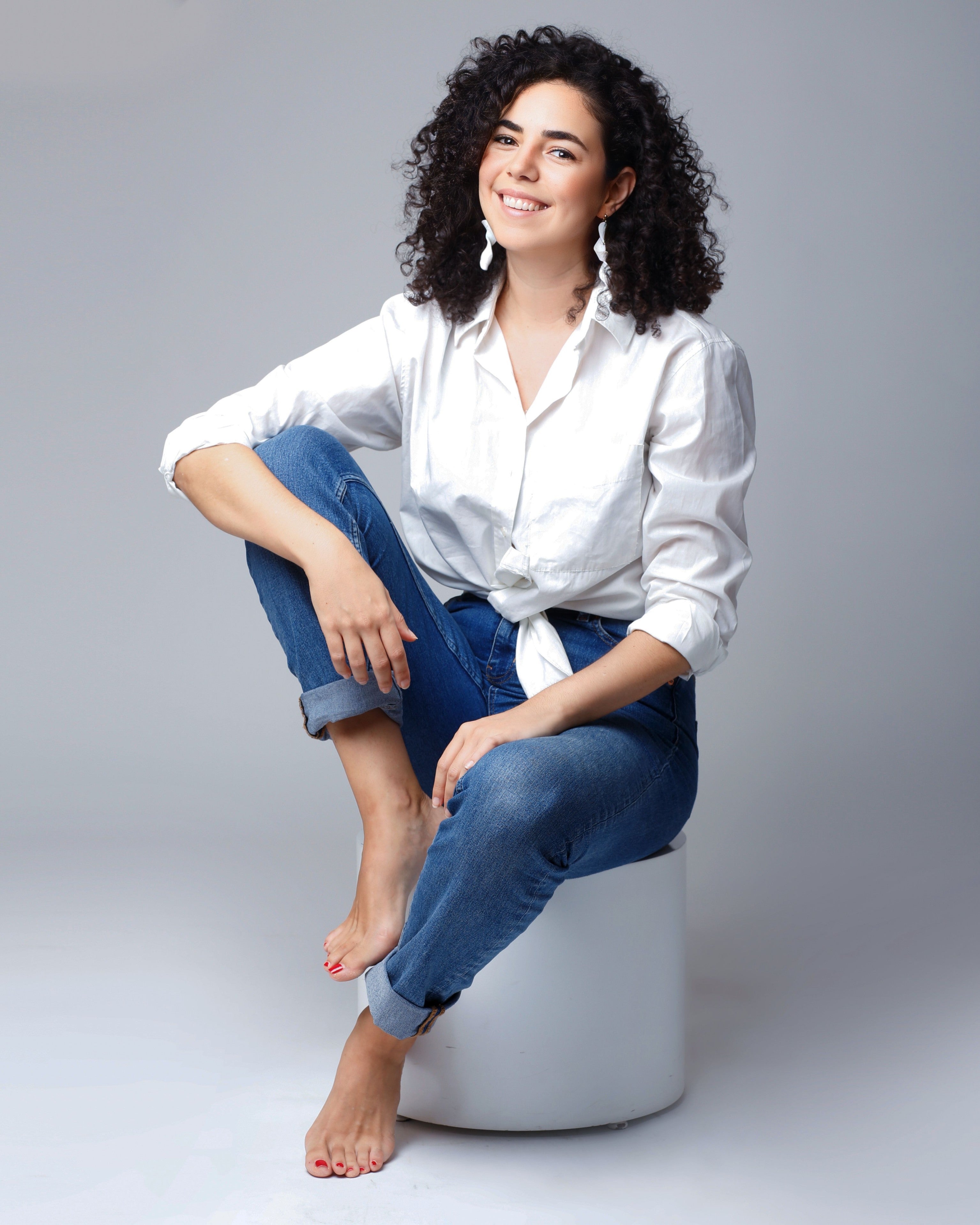 Kenza sitting on a white stool wearing a white shirt and blue jeans against a gray background